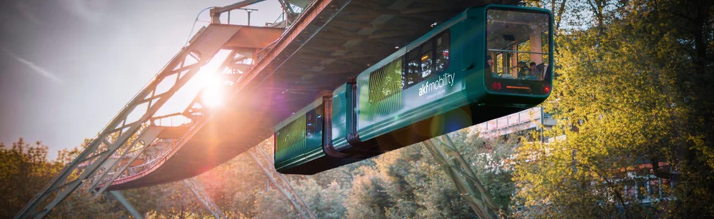 Schwebebahn mit zwei Waggons fährt auf einer Stahlkonstruktion bei Sonnenuntergang über bewaldetes Gelände